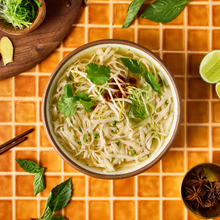 Load image into Gallery viewer, A top-down view of a bowl of noodle soup filled with bean sprouts, cilantro, and chili oil, set on a tiled orange surface with lime wedges, herbs, and chopsticks around it.
