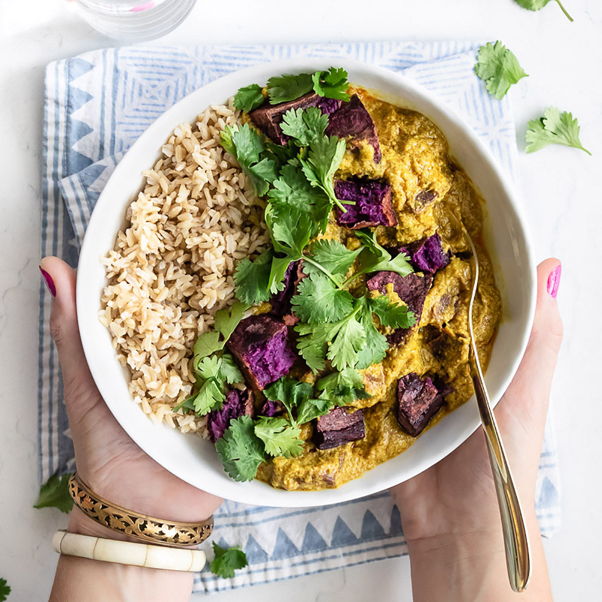 Hands holding a white ceramic bowl of purple sweet potato curry with brown rice. A fork is in the food.