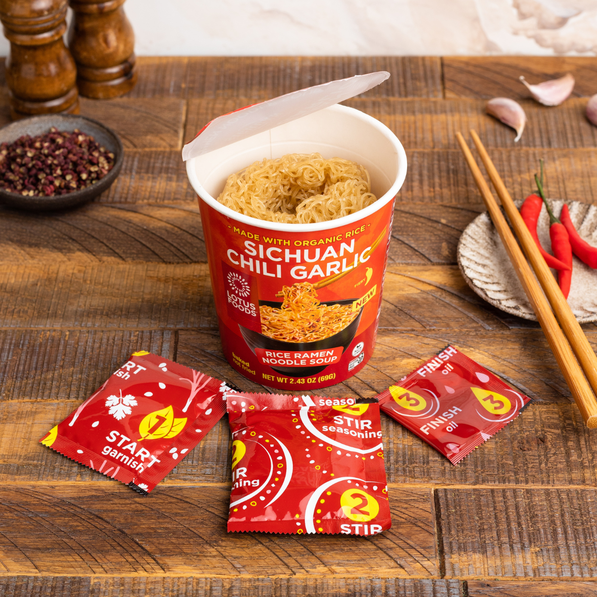 An open Lotus Foods Sichuan Chili Garlic Rice Ramen Noodle Soup Cup, noodles visible in cup, seasoning packets and chopsticks alongside, on a dark wooden backdrop.