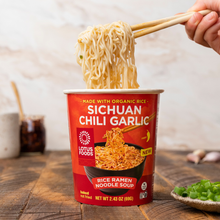 Load image into Gallery viewer, A hand pulling noodles from a cup of Lotus Foods Sichuan Chili Garlic Rice Ramen Noodle Soup, shown in its red packaging against a dark wooden backdrop.

