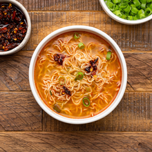 Load image into Gallery viewer, Overhead shot of a white ceramic bowl of Lotus Foods Sichuan Chili Garlic Rice Ramen Noodle Soup, on a dark wooden backdrop.
