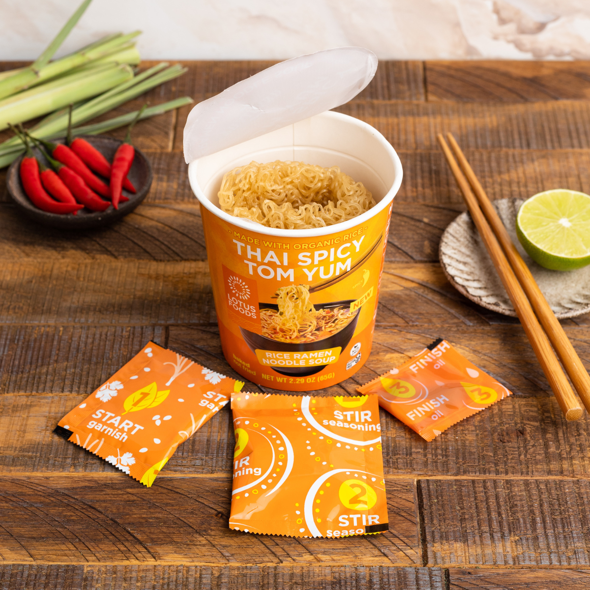 An open Lotus Foods Thai Spicy Tom Yum Noodle Soup Cup, noodles visible in cup, seasoning packets and chopsticks alongside, on a dark wooden backdrop.