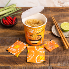Load image into Gallery viewer, An open Lotus Foods Thai Spicy Tom Yum Noodle Soup Cup, noodles visible in cup, seasoning packets and chopsticks alongside, on a dark wooden backdrop.
