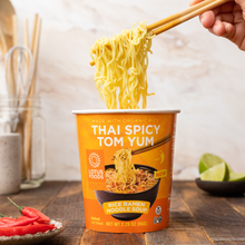 Load image into Gallery viewer, A hand pulling noodles from a cup of Lotus Foods Thai Spicy Tom Yum Rice Ramen Noodle Soup, shown in its red packaging against a dark wooden backdrop.
