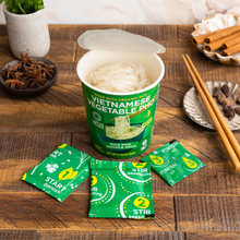 Load image into Gallery viewer, An open Lotus Foods Vietnamese Vegetable Pho Noodle Soup Cup, noodles visible in cup, seasoning packets and chopsticks alongside, on a dark wooden backdrop.
