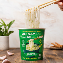 Load image into Gallery viewer, A hand pulling noodles from a Lotus Foods Vietnamese Vegetable Pho Noodle Soup Cup on a dark wooden backdrop.
